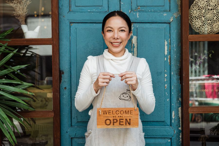 A smiling café owner holding a welcome sign at her shop door